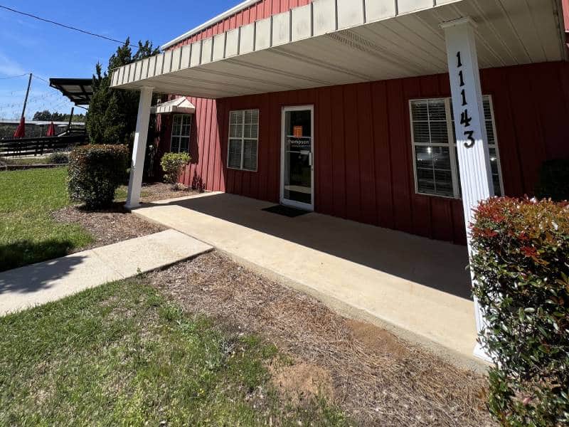 Red building with white columns and sidewalk entrance surrounded by greenery under a clear blue sky.