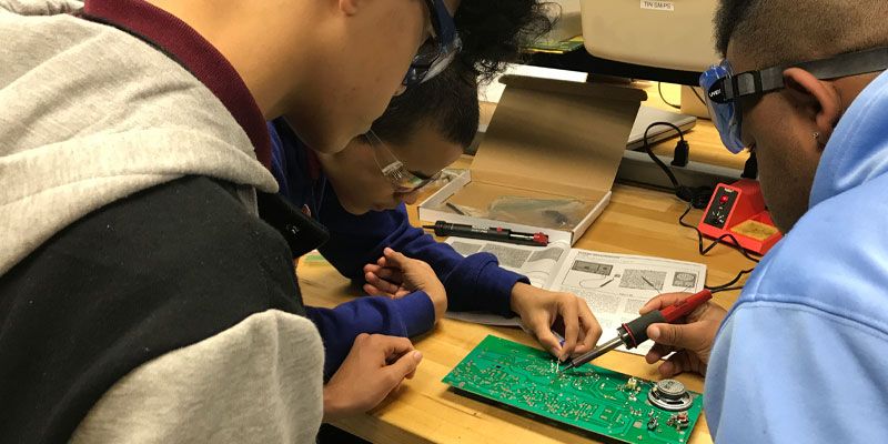 Students collaborating on a circuit board project in a classroom setting, wearing safety goggles for protection.