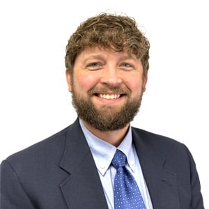Man in a suit smiling confidently against a white background, wearing a blue tie and curly hair. Professional headshot.
