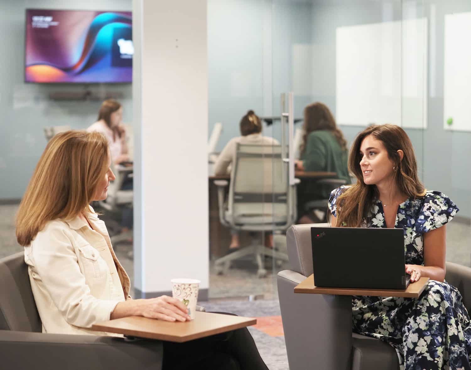 Two women having a conversation in a modern office lounge with others working in the background.