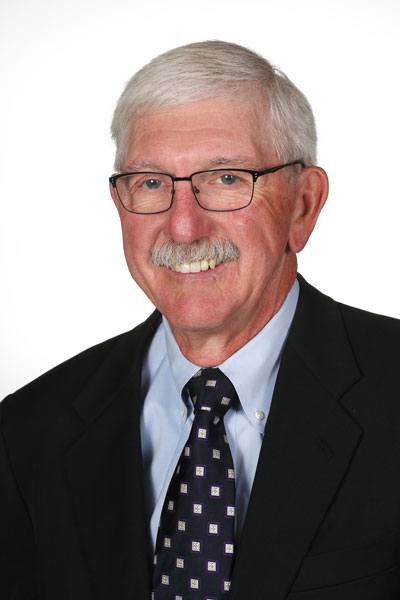 Elderly man in suit smiling, wearing glasses, gray hair, mustache, white background.