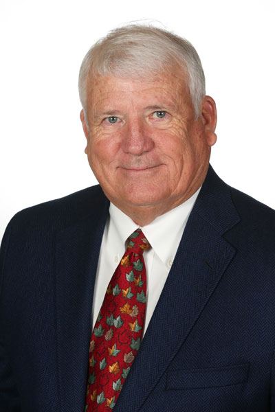 Elderly man in a suit with a white shirt and patterned tie, smiling against a white background.