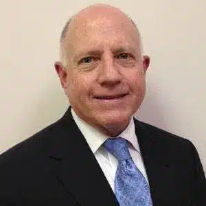 Man in a suit with a blue tie, smiling in front of a plain background.