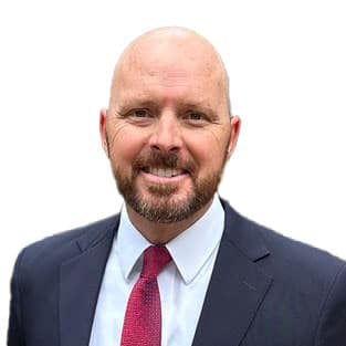 A man with a beard in a suit and tie smiles against a white background.