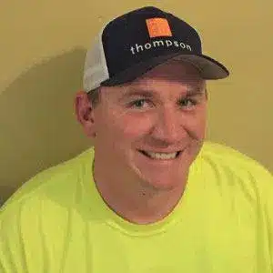 Man in a yellow shirt wearing a black and white thompson cap, smiling in front of a beige background.