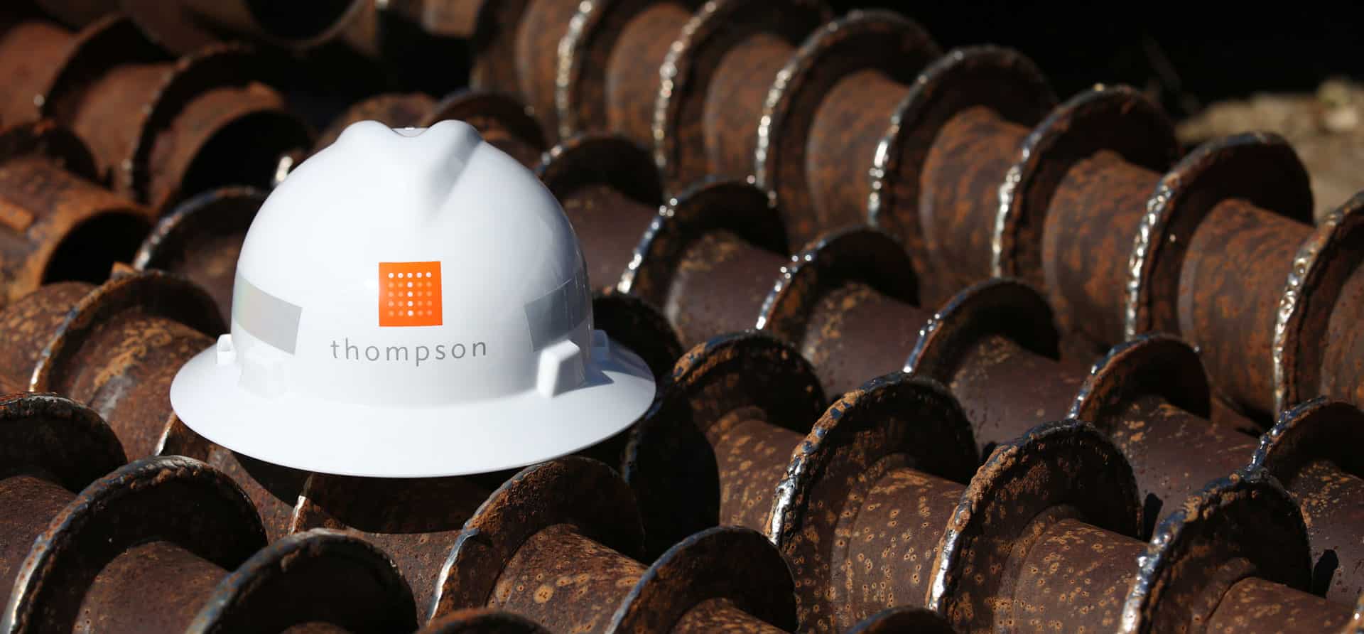 White hard hat with Thompson logo on rusty industrial drill bits background.