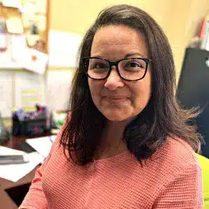 Woman wearing glasses and a pink sweater, smiling in an office setting with papers and books in the background.